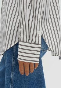 Weißes Hemd mit schmalen schwarzen Streifen, geknöpften Manschetten und einer Taschenapplikation, kombiniert mit blauen Jeans aus Denim.