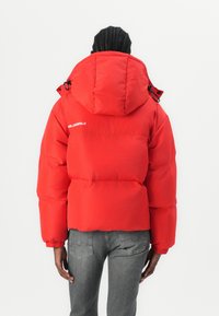 Red puffer jacket with a hood, featuring a front zip closure and elastic cuffs. Back displays logo text and a quilted design.