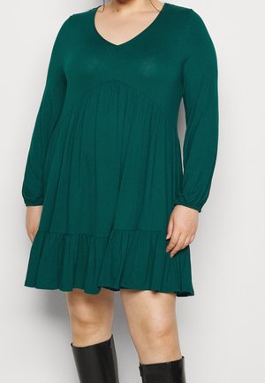 Vrouw draagt een donkergroene V-halsjurk met lange mouwen en geplooide tiers, gecombineerd met zwarte kniehoge laarzen, staand tegen een witte achtergrond.