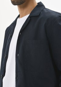 Camisa de botones azul marino de tejido ligero, con cuello, mangas cortas y un bolsillo de parche en el lado izquierdo.