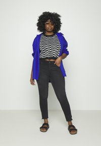 Modelo vestindo um suéter cropped com padrão ondulado em preto e branco, camisa azul, jeans skinny pretos e sandálias pretas com fivela.