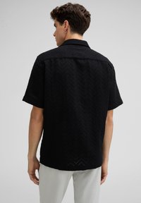 Schwarzes Kurzarmhemd mit strukturiertem Zickzackmuster, mit einem Kragen und geradem Saum hinten, getragen über hellen Hosen.