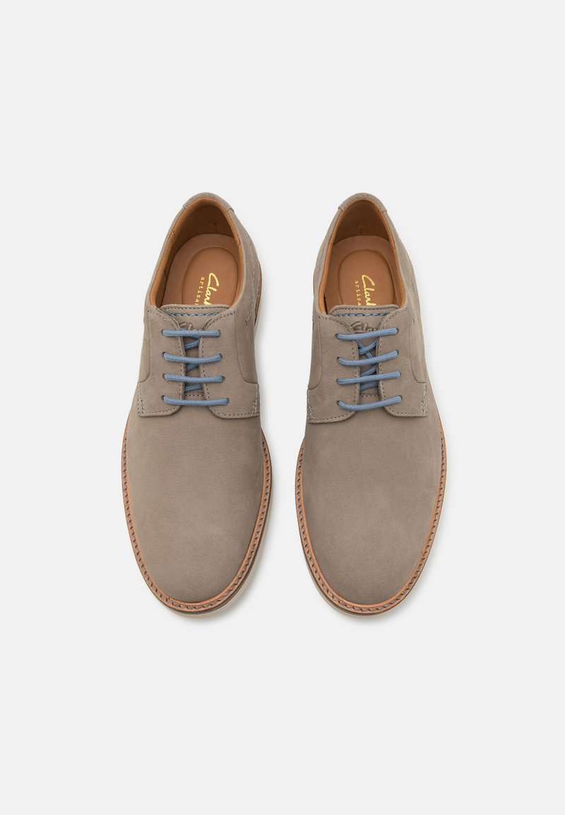 Zapatos de ante grises con cordones azules, punta redondeada y suela de cuero cosida en tono marrón. Textura suave y diseño minimalista con logo de la marca grabado en el interior.
