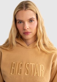 Kapučiukas švelnios béžinės spalvos su iškilniu "BIG STAR" logotipu, kuris dera su spalva, pasižymintis laisvu siluetu ir tekstūruotu audiniu.