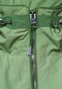 Ljusgrön jacka med insidan som har en metallisk dragkedja, flätade dragsnören med lila spetsar och en slät, lätt rynkad tygtextur.