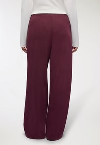 Bordowe spodnie szerokie o gładkiej fakturze i elastycznym pasie, pokazane z tyłu, w połączeniu z dopasowaną białą koszulą z długim rękawem.
