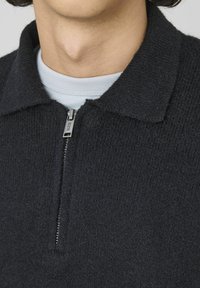 Pullover noir en tricot avec un zip demi-long et un col. Texture unie et détail en zip argenté. Porté par-dessus un t-shirt col rond bleu clair.