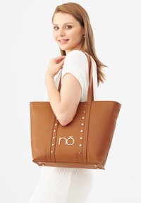 Braune Leder-Tote mit silberner Hardware, die vertikale Streifen und dekorative Nieten aufweist. Die Front zeigt das Logo "nō". Glatte Textur, breite Form.