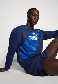 Felpa blu navy con un grande logo Puma blu, colletto a girocollo e maniche lunghe, abbinata a pantaloni corti blu navy. Texture liscia.