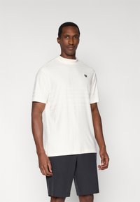 Biały sportowy T-shirt z krótkim rękawem, wysokim kołnierzem, teksturowanym wzorem i małym logo na piersi. Noszony z ciemnymi szortami.