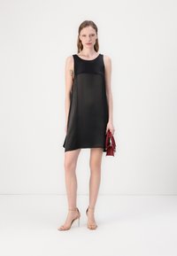 Rochie fără mâneci din mătase neagră, cu o croială lejeră, cu hem la nivelul genunchilor, accesorizată cu o geantă roșie și sandale cu toc nude.