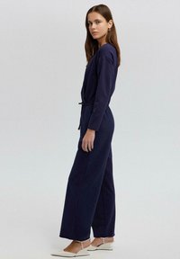 Námornícky modrý overal s dlhými rukávmi, sťahovacím pásom a širokým strihom nohavíc. Hladká textúra, minimálne detaily a elegantný silueta.