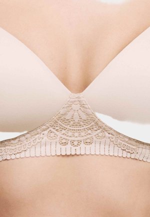 Béžová podprsenka s tvarovanými košíčky a složitým krajkovým zdobením pod prsy, zobrazena na horní části hrudi a zádech osoby.