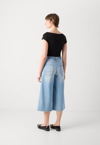 Svart tettsittende topp, lyseblå vide denim culotter med høy midje og baklommer, samt svarte flate sko, sett fra baksiden.