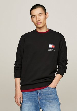 Sudadera negra con cuello redondo, que presenta un parche de logo multicolor en el pecho, superpuesta a una camiseta color burdeos, llevada con jeans azules.