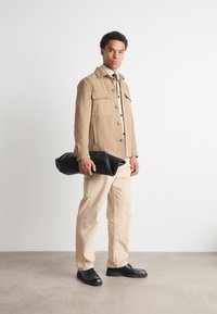 Veste en coton beige avec poches frontales, boutons noirs et col à rabat. Pantalons beiges et chaussures noires. Tient un sac noir roulé.