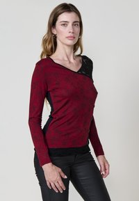 Červený long-sleeve top s kvetinovým vzorom, čiernym čipkovaným výstrihom a bočnými panelmi, so zapínaním na gombíky dole a priliehavým strihom.
