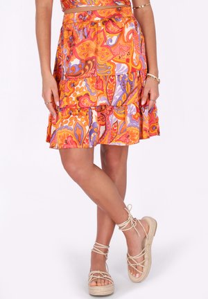 Vrouw draagt een kleurrijke oranje, roze en paarse bloemenrok en beige espadrille sandalen met veters en enkelbandjes, staand met gekruiste benen.