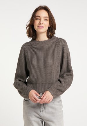 Ung kvinde med skulderlangt brunt hår iført en grå striktrøje og lysegrå jeans, der smiler let med hænderne foldet.