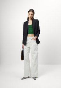 Blazer nero su misura, top verde corto, pantaloni bianchi a gamba larga e tacchi con stampa zebra. Tiene in mano una borsa nera e gialla. Sfondo minimalista.