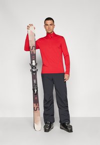 Pull rouge à manches longues avec une fermeture éclair sur le devant, assorti à un pantalon de ski noir et des chaussures de ski noires, tenant des skis marron et noir avec fixations.