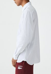 Cămașă albă cu mâneci lungi, având un guler clasic, manșete cu nasturi și un luciu subtil, asortată cu pantaloni de trening VIȘINE cu broderie logo.