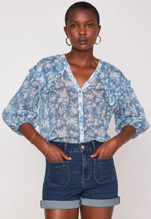 Donna che indossa una blusa blu a fiori con bottoni e maniche a sbuffo, pantaloni corti di denim scuro arrotolati, mani nelle tasche, orecchini a cerchio dorati e braccialetti.