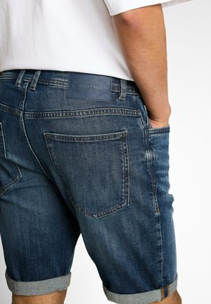 Man wearing rolled-up blue denim shorts with back pockets and a white shirt, hand in front pocket, against a plain background.