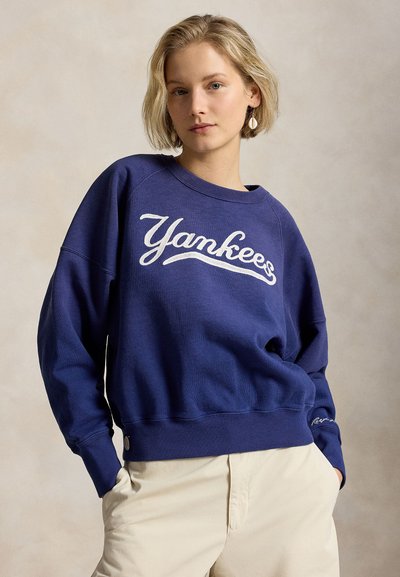 Jeune femme aux cheveux blonds courts portant un sweat-shirt bleu "Yankees" et un pantalon beige, debout les mains dans les poches devant un fond uni.
