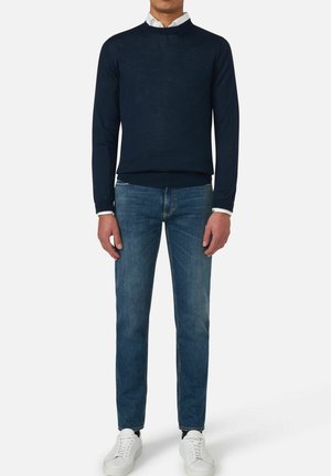Marineblaues Strickpullover mit Rundhalsausschnitt, kombiniert mit mittelblauen Jeans. Das Model trägt weiße Sneaker. Glatte Textur, lässiges Design.