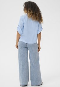 Blouse à manches courtes, bleu clair, à boutons avec des poignets retroussés, associée à un jean en denim clair à jambes larges. Tissu lisse et coupe décontractée.