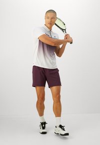 Tenis odijelo koje uključuje gradijent bijelu i ljubičastu polo majicu, bordo šortse i crno-bijele teniske tenisice s čarapama doankla. Drži reketa.