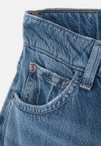 Jasnoniebieskie dżinsy z wysokim stanem, wyhaftowanymi kieszeniami i małym dekoracyjnym akcentem przy przedniej kieszeni.