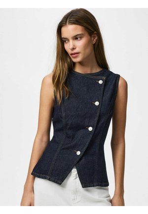Kvinde iført mørk denim top uden ærmer med hvide knapper fastgjort asymmetrisk, kombineret med hvide bukser, mod en ensfarvet baggrund.