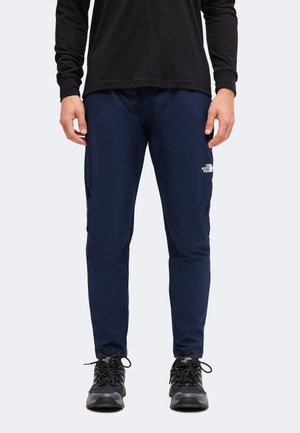Man draagt marineblauwe The North Face-broek en zwart shirt met lange mouwen met zwarte wandelschoenen tegen een witte achtergrond.