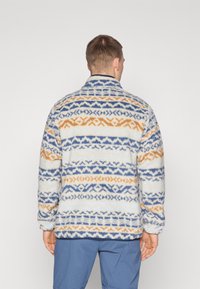 Φλις ζακέτα σε κρεμ απόχρωση με μπλε και πορτοκαλί γεωμετρικά σχέδια. Κανονικός γιακάς, μακριά μανίκια και ελαστικές μανσέτες. Μαλακό, υφασμένο ύφασμα.