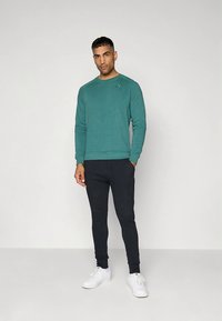 Felpa color teal con scollo rotondo, abbinata a pantaloni jogger neri. L'outfit presenta un tessuto testurizzato e scarpe da ginnastica bianche. Dettaglio sottile con logo.