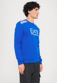 Modrá mikina so štruktúrovaným povrchom, s bielym logom "EAT EMPORIO ARMANI" a pruhovanými detailmi na ramenách. Výstrih do kruhu a dlhé rukávy.