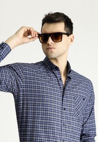 Camicia a righe check blu e bianca con taschino e maniche arrotolate, abbinata a occhiali da sole con tonalità marroni. Tessuto dalla trama liscia.