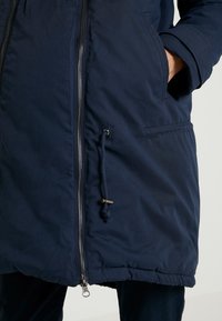 Zbliżenie osoby noszącej granatową parkę z widocznym srebrnym zamkiem błyskawicznym i ręką włożoną do bocznej kieszeni.