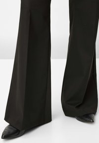 Zwarte wijde broek van gladde stof. Heeft een hoge taille en een brede pijp, met puntige zwarte schoenen die onder de zoom vandaan gluren.