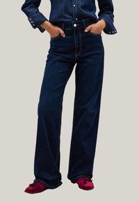 Pantalones de mezclilla de pierna ancha, de color azul oscuro y de cintura alta, con bolsillos frontales y detalles de costura visibles. Llevados con calzado suave de color rojo.
