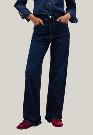 Pantaloni di jeans blu scuro, a gamba larga, con vita alta, tasche frontali e dettagli di cucitura visibili. Indossati con scarpe morbide rosse.