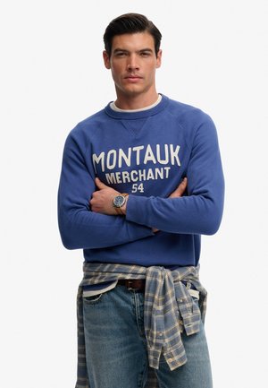 Man met donker haar, gekleed in een blauwe "Montauk Merchant 54" sweatshirt, geruite blouse om de taille geknoopt, spijkerbroek en een horloge om de pols, met gekruiste armen.