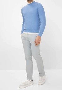 Man in een lichtblauwe trui, witte onderhemd, lichtgrijze broek en witte sneakers, staande met zijn handen in de zakken.