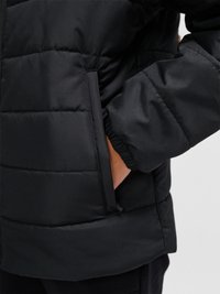 Hand in der Tasche einer schwarzen, gesteppten Daunenjacke mit elastischem Ärmelbündchen, Vorderseite und Steppdetails sichtbar.