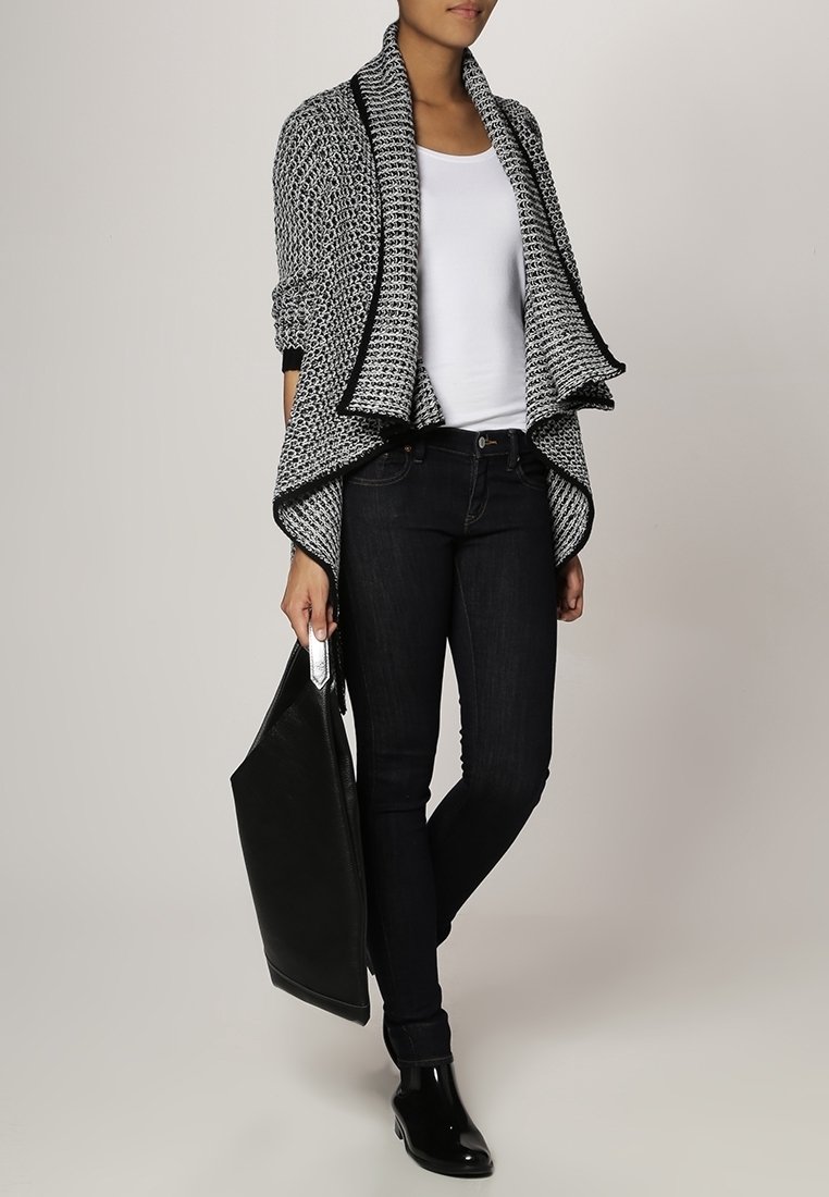 Cárdigan de punto en blanco y negro con cuello de chal, llevado sobre una blusa blanca. Jeans oscuros y botas de tobillo brillantes. Se presenta un gran bolso tote negro.