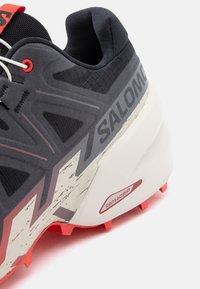 Μαύρο αθλητικό παπούτσι με πλέγμα στην επάνω πλευρά και κόκκινες λεπτομέρειες. Διαθέτει αντίθετη εξωτερική σόλα, υφασμένες πλαϊνές επιφάνειες και επιβλητικό λογότυπο της μάρκας.