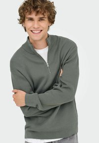 Suéter verde con canalé y cuello con cremallera, textura suave, corte relajado, llevado sobre una camiseta blanca, contra un fondo liso.