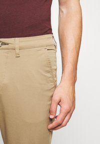 Tan chinos met een gladde textuur, voorzien van zijzakken, zichtbare riemlussen en een platte tailleband. Hand gepositioneerd nabij het been.